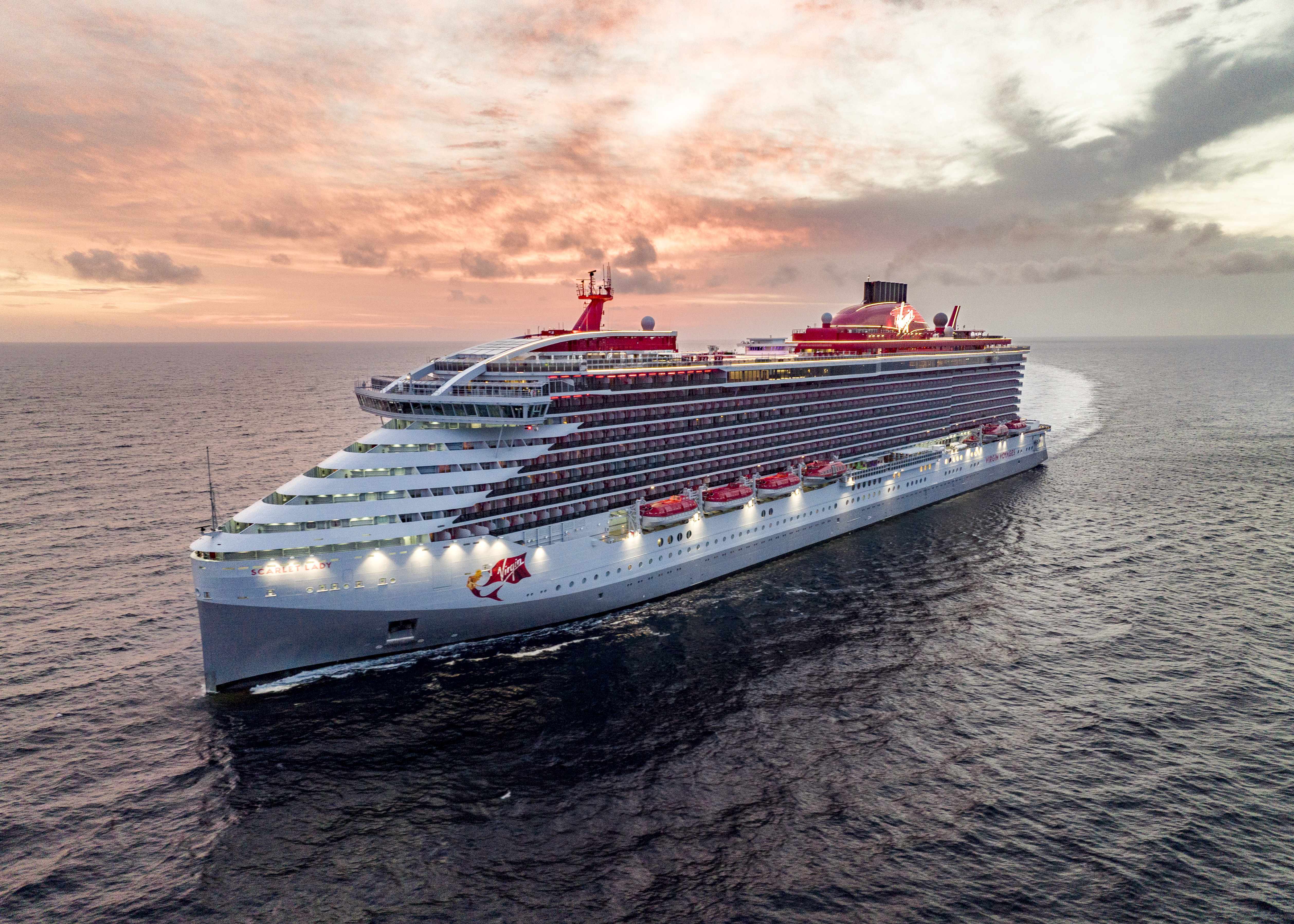 Virgin Voyages Scarlet Lady cruise ship with red mermaid tail logo, sailing on calm ocean waters during golden hour with dramatic sky